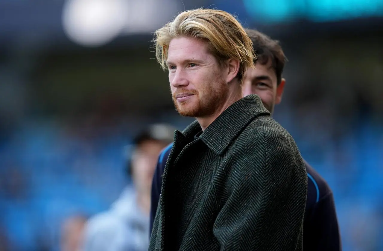 Kevin De Bruyne, wearing a coat, looks on from pitchside at the Etihad Stadium