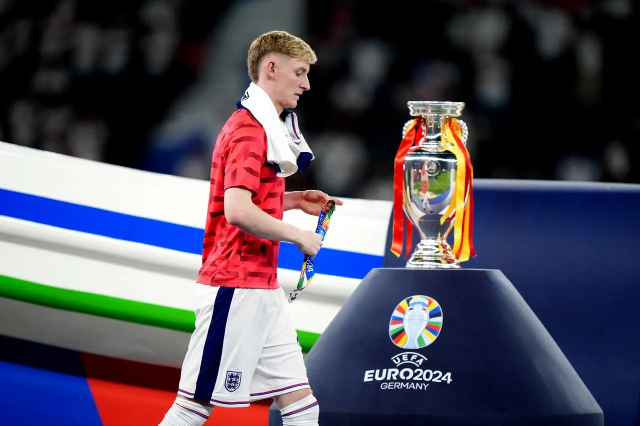 Anthony Gordon, with a shirt round his neck, walks past the Euro 2024 trophy