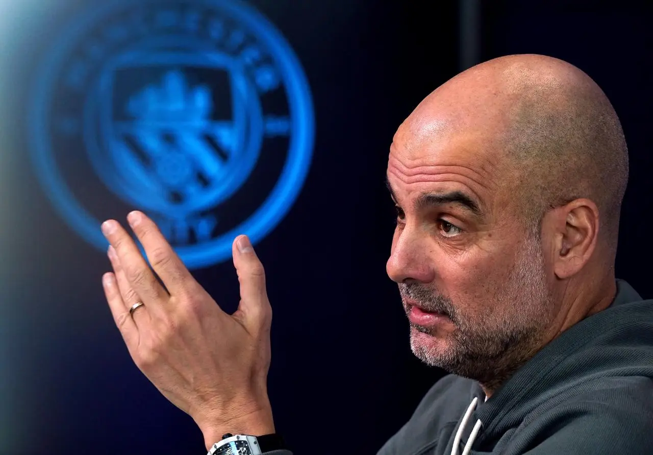 Pep Guadiola gestures while sitting in front of a Manchester City crest