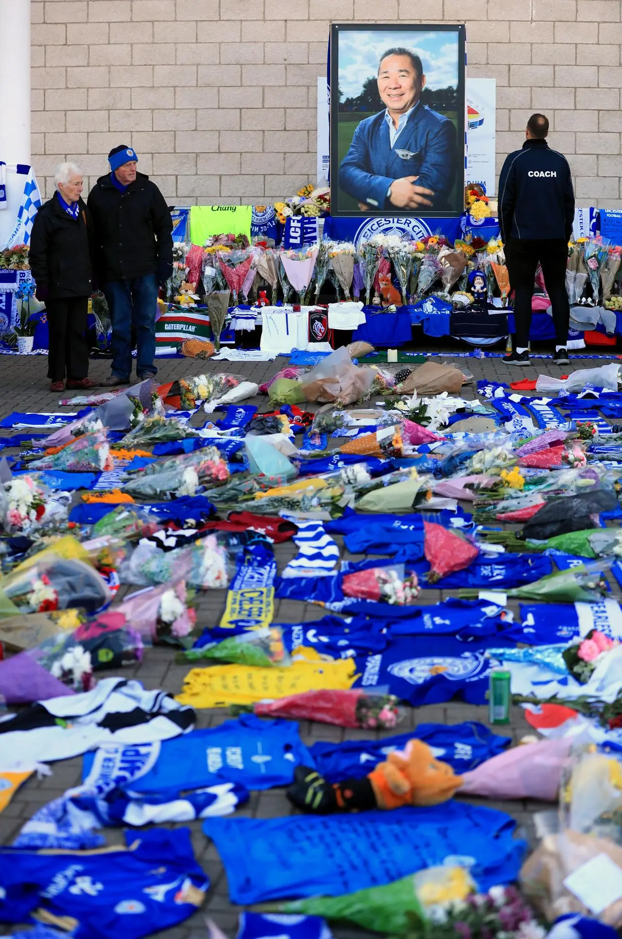 People look over tributes of flowers, blue football shirts and stuffed teddies that cover the floor and along the wall where a portrait of Vichai Srivaddhanaprabha stands